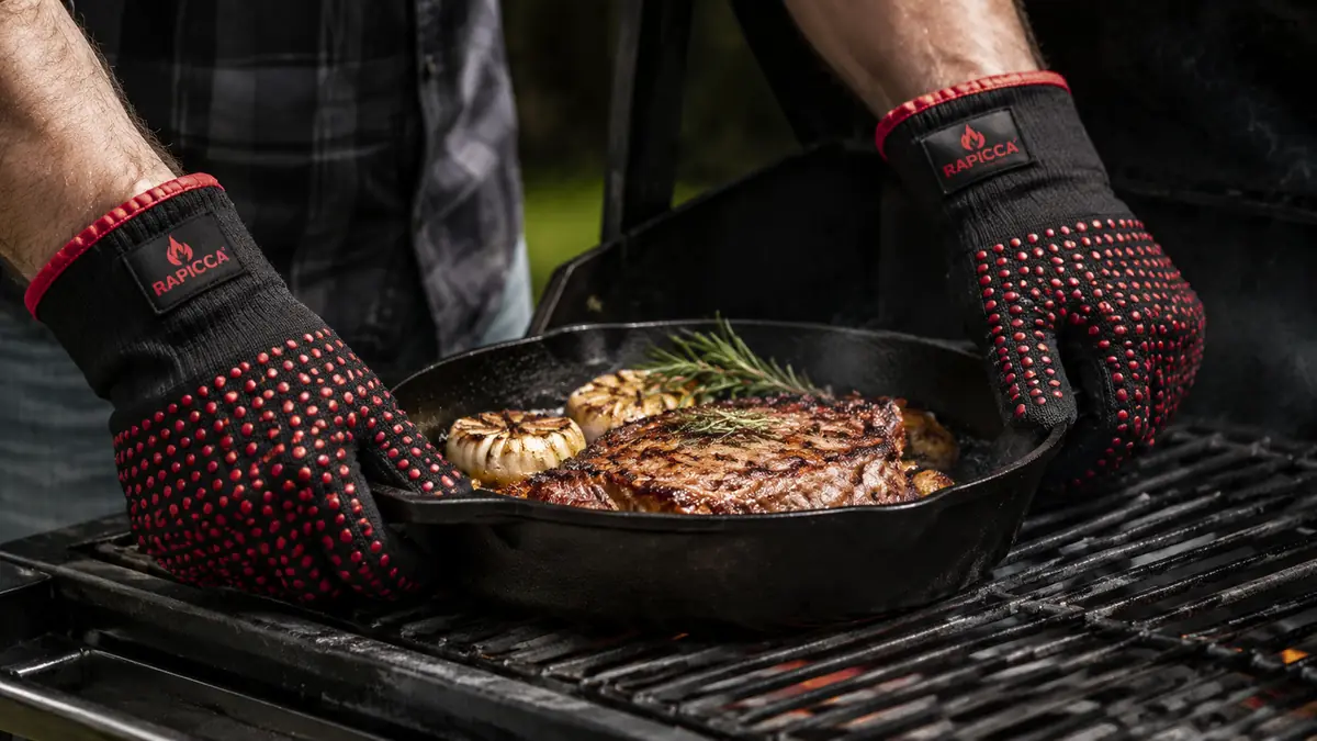 Close-up of hands in grill gloves gripping a hot cast iron pan straight off the grill grate