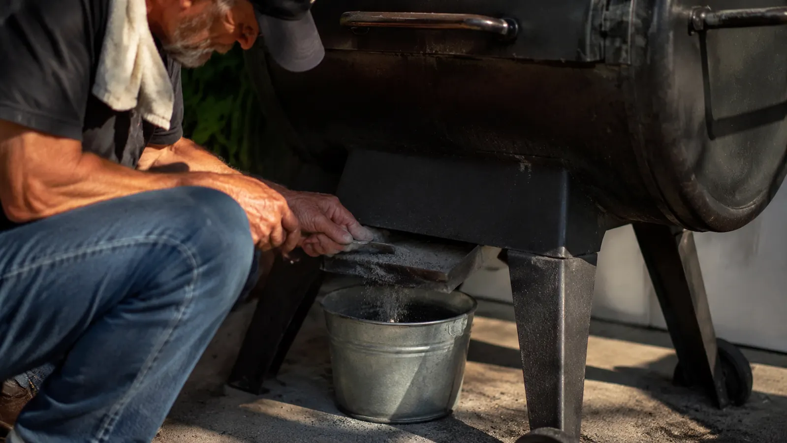 Close-up of a hand emptying gray wood pellet ash from the burn pot area of a Pit Boss grill into a metal bucket
